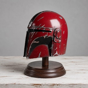 Op de afbeelding: Een rood en zwart helm met zilveren accenten, gepresenteerd op een houten standaard. De helm heeft een verweerde uitstraling, wat duidt op schade. De standaard is donkerbruin en de achtergrond is neutraal grijs.