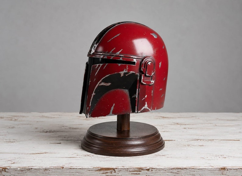 Op de afbeelding: Een rood en zwart helm met zilveren accenten, gepresenteerd op een houten standaard. De helm heeft een verweerde uitstraling, wat duidt op schade. De standaard is donkerbruin en de achtergrond is neutraal grijs.
