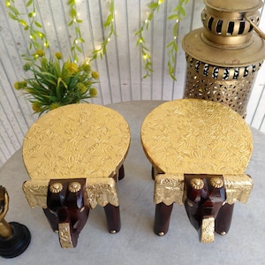 Puede incluir: Dos taburetes decorativos de madera con forma de elefante. Cada taburete tiene una parte superior dorada con estampado floral y patas de color marrón oscuro. Las patas delanteras con forma de elefante están adornadas con detalles dorados. Los taburetes miden unos 15 cm de altura.