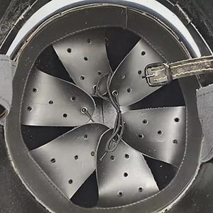 Op de afbeelding: Binnenaanzicht van een zwarte helm met een leren rand en een reeks metalen platen. De platen zijn geklonken en geperforeerd met kleine gaatjes. Een leren riem met een gesp is zichtbaar.
