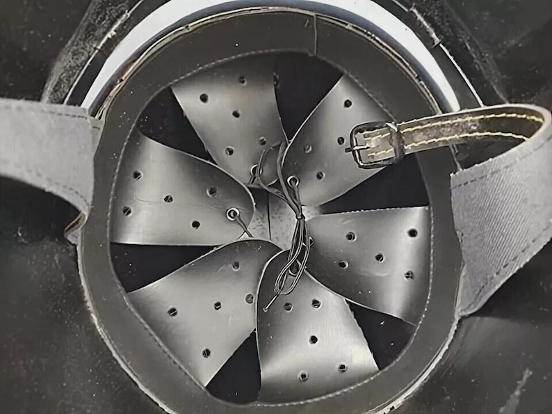 Op de afbeelding: Binnenaanzicht van een zwarte helm met een leren rand en een reeks metalen platen. De platen zijn geklonken en geperforeerd met kleine gaatjes. Een leren riem met een gesp is zichtbaar.