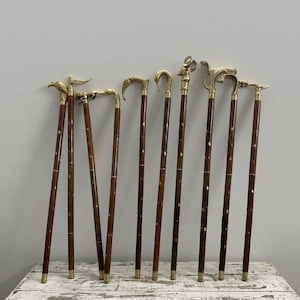 May include: A collection of antique walking canes with ornate brass handles. The dark brown wooden shafts are inlaid with small, decorative accents. Each cane features a unique handle design, including bird, animal, and curved shapes. The canes are displayed on a distressed white wooden surface.