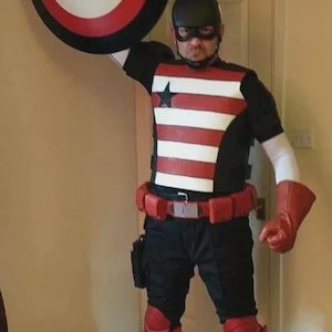 Puede incluir: Un disfraz de superh&eacute;roe con un escudo. El disfraz incluye una parte superior en blanco y negro con rayas rojas, un cintur&oacute;n rojo y botas rojas. El escudo es circular con c&iacute;rculos conc&eacute;ntricos rojos, blancos y negros.