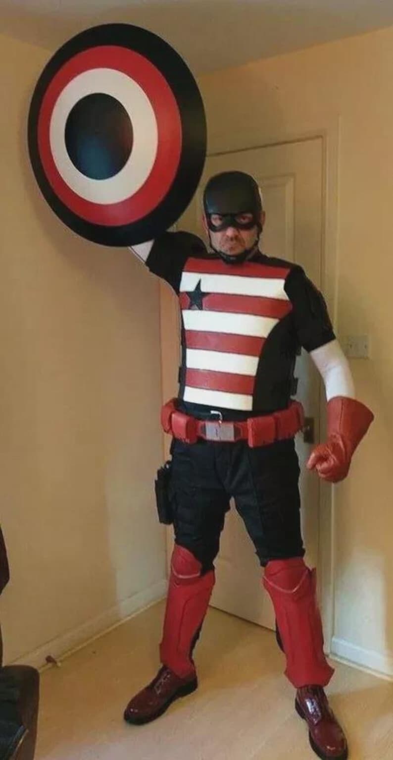 Puede incluir: Un disfraz de superh&eacute;roe con un escudo. El disfraz incluye una parte superior en blanco y negro con rayas rojas, un cintur&oacute;n rojo y botas rojas. El escudo es circular con c&iacute;rculos conc&eacute;ntricos rojos, blancos y negros.