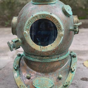 Könnte beinhalten: Antiker Taucherhelm mit Kupfer- und Grünspan-Finish. Der Helm hat ein rundes Sichtfenster und den Text "ANCHOR ENGINEERING 1921". Der Helm hat mehrere Anschlüsse und eine Grundplatte.
