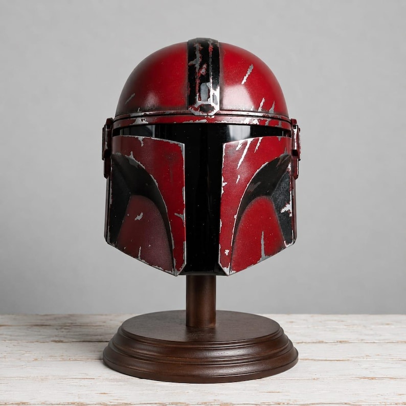 Op de afbeelding: Een rood en zwart Mandalorian helm met een verweerde afwerking, tentoongesteld op een houten standaard. De helm heeft een zwart vizier en zilveren accenten, met een door de strijd geteisterde uitstraling. De standaard is donkerbruin.