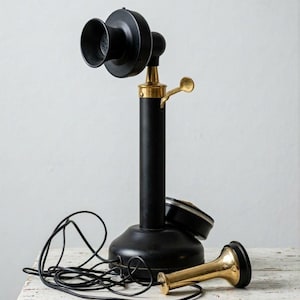 May include: A vintage black candlestick telephone with brass detailing. The phone features a round base, a central column, and a large earpiece. The handset is separate, and a black cord connects the phone.