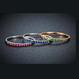 May include: Three delicate rings with a row of gemstones. One ring features red gemstones, another has blue, and the third has green. The rings are set against a dark background, showcasing the colors and details of the jewelry.