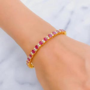 May include: A gold bracelet featuring alternating oval-cut red gemstones and small white gemstones. The bracelet is worn on a wrist, showcasing the jewelry's design and color contrast.