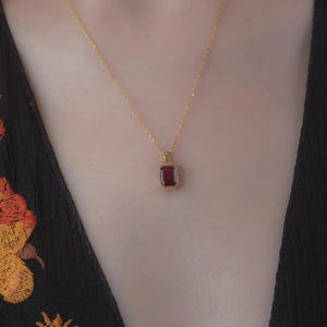 Puede incluir: Un collar dorado con un colgante rectangular de piedra preciosa rojo oscuro. El colgante está engastado en un marco dorado y cuelga de una delicada cadena dorada. El collar se muestra sobre un fondo oscuro.