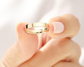 May include: A gold wedding band with a flat, polished surface. The ring is held between two fingers, showcasing its simple, elegant design. The band's color is a bright, warm yellow, reflecting light.