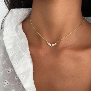 May include: A delicate gold-toned necklace featuring a pair of small, sparkling wing pendants. The wings are adorned with tiny, clear gemstones. The necklace is worn on a person's neck, against a white shirt.