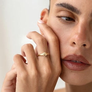 Puede incluir: Un anillo de oro con un pequeño diseño de concha marina en relieve en la banda. El anillo tiene un acabado metálico pulido y una banda delgada y cónica que se asienta en el dedo, mostrando un motivo de concha en forma de abanico detallado.
