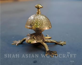 May include: A brass frog-shaped bell with a floral patterned dome and a small handle. The bell is resting on a gray surface. The text "SHAH ASIAN WOODCRAFT" is visible on the base.