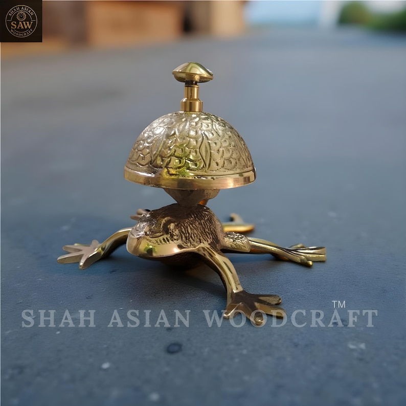 May include: A brass frog-shaped bell with a floral patterned dome and a small handle. The bell is resting on a gray surface. The text "SHAH ASIAN WOODCRAFT" is visible on the base.