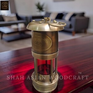 Op de afbeelding: Een vintage messing mijnwerkerslamp met een rode glazen behuizing. De lamp heeft een handvat, een decoratief ovaal plaatje met tekst en de woorden "LONDON 1920". De basis en de bovenkant zijn cirkelvormig. Van Shah Asian Woodcraft.
