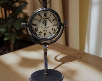 May include: A vintage-style clock with a round face and Roman numerals. The clock face reads "Victoria London 1876". The clock is mounted on a dark metal stand with a circular base. The clock is a decorative item.