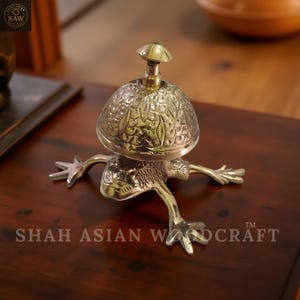 May include: A brass frog-shaped bell with a mushroom-shaped top. The bell has intricate leaf-like patterns and is set on a dark wooden surface. The text "SHAH ASIAN WOODCRAFT" is visible.