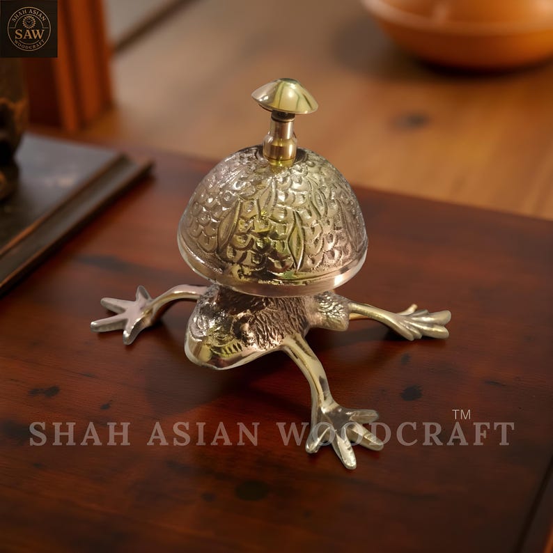 May include: A brass frog-shaped bell with a mushroom-shaped top. The bell has intricate leaf-like patterns and is set on a dark wooden surface. The text "SHAH ASIAN WOODCRAFT" is visible.