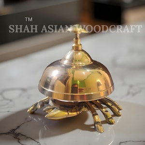 Op de afbeelding: Een gepolijste messing bel met een krabachtig onderstel. De bel heeft een koepelvorm met een klein handvat aan de bovenkant. De basis heeft meerdere krabbenpoten. De tekst "SHAH ASIAN WOODCRAFT" is zichtbaar.