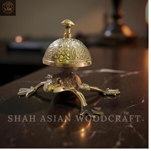 May include: A brass frog-shaped bell with a dome-shaped top. The bell has an ornate floral design and a small handle. The base of the bell is a detailed frog with outstretched legs. The text "SHAH ASIAN WOODCRAFT" is visible.
