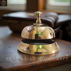 May include: A polished brass bell with a rounded top and a flared base. The bell has a small, decorative handle on top. The words "SHAH ASIAN WOODCRAFT" are printed on the base of the bell. The bell is sitting on a wooden surface.