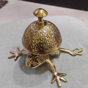 May include: A brass frog-shaped bell with a mushroom-shaped handle. The bell has an ornate, textured surface. The frog's legs are outstretched, giving the bell a whimsical appearance. The bell is made of shiny, polished brass.