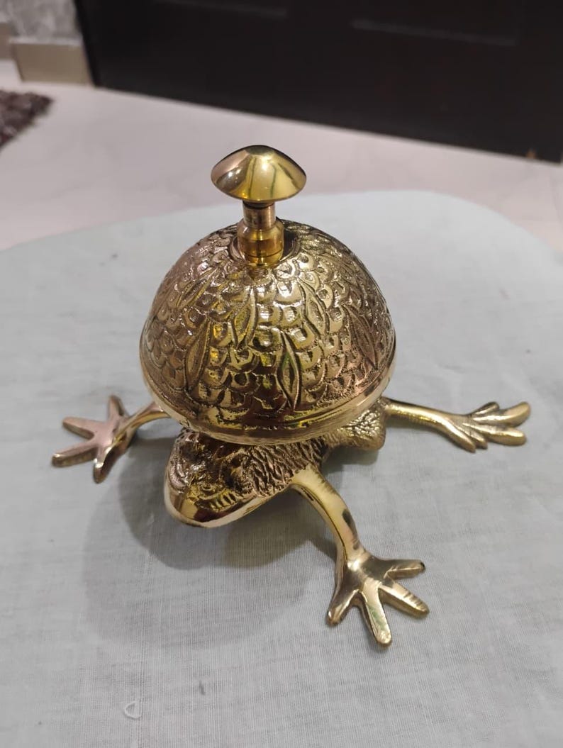 May include: A brass frog-shaped bell with a mushroom-shaped handle. The bell has an ornate, textured surface. The frog's legs are outstretched, giving the bell a whimsical appearance. The bell is made of shiny, polished brass.
