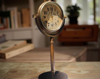 May include: A vintage-style brass desk clock with a round face. The clock has a cream-colored dial with black numerals and hands. The clock is supported by a slender, vertical stand and a circular base. The clock's face is approximately 4 inches in diameter.