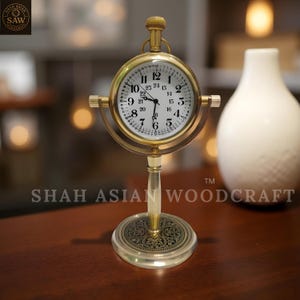 May include: A brass-colored, vintage-style clock with a white face and black numerals. The clock is mounted on a decorative stand. The clock's face displays the time. The base of the clock has an ornate design. The clock is approximately 6 inches tall.