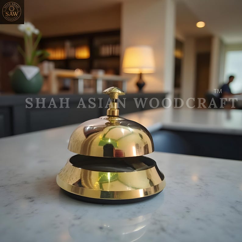 May include: A polished brass service bell with a rounded top and a flared base. The bell has a small, decorative handle on top. The text "SHAH ASIAN WOODCRAFT" is visible. The bell is sitting on a white marble surface.