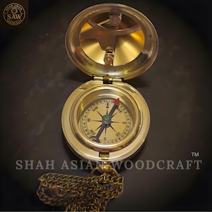 May include: A brass compass with a hinged lid, open to reveal a compass dial with directional markings. The compass has a chain attached. The text "SHAH ASIAN WOODCRAFT" is visible.