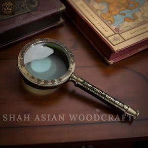May include: A vintage brass magnifying glass with a dark handle, resting on a wooden surface. The magnifying glass has a circular lens and decorative detailing around the rim. The handle is engraved with text.