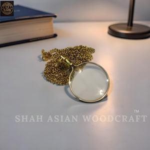May include: A gold-coloured magnifying glass with a chain, lying on a white surface. The magnifying glass has a clear lens and a decorative handle. The chain is made of small, interlocking links. The text "SHAH ASIAN WOODCRAFT" is visible.
