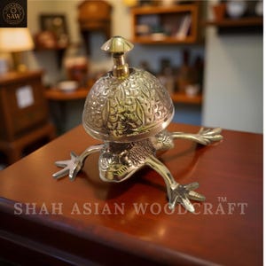 May include: A brass frog-shaped bell with a mushroom-shaped top. The bell has intricate floral patterns and is sitting on a wooden surface. The text "SHAH ASIAN WOODCRAFT" is visible on the surface.