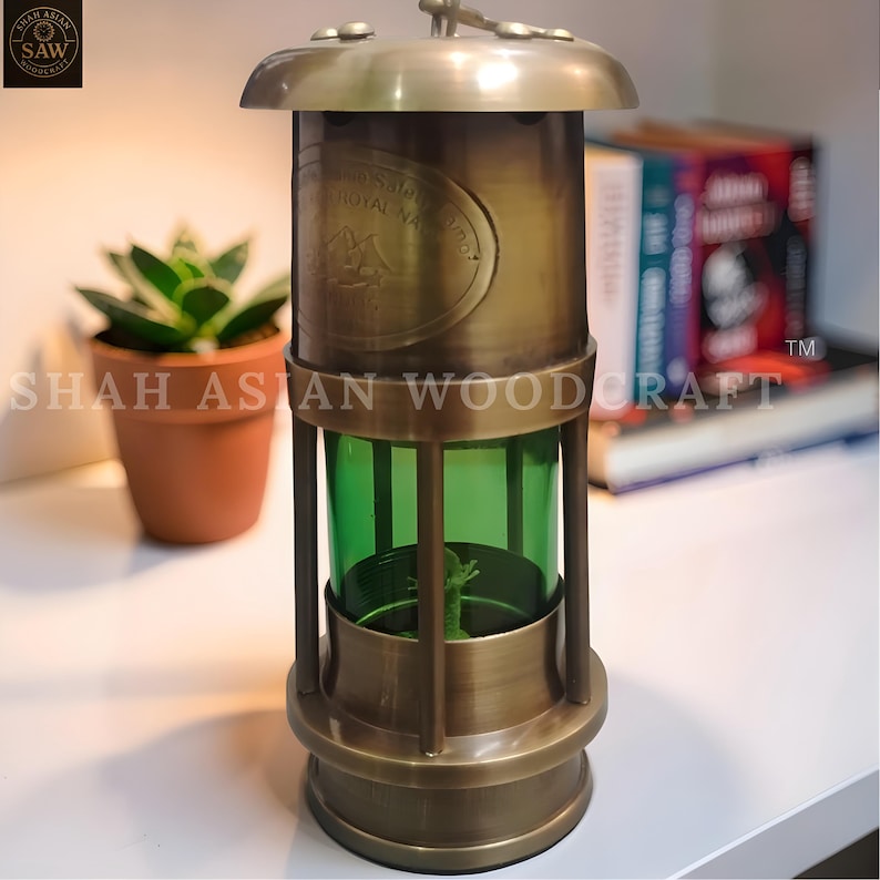 Op de afbeelding: Een antieke messingkleurige lantaarn met een groene glazen binnenkant. De lantaarn heeft een ronde boven- en onderkant, met verticale staven en een handvat. De tekst "Royal Navy" is zichtbaar. Een kleine plant in een pot staat op de achtergrond.