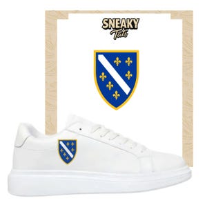 Könnte beinhalten: Weiße Sneaker mit niedrigem Profil. Die Schuhe haben ein Schildemblem mit blauem und gelbem Design und einem weißen Streifen. Das Bild enthält auch eine Grafik mit dem Text "Sneaky Tats".