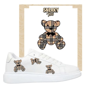 May include: White trainers with a teddy bear design. The trainers feature a repeating pattern of teddy bears in a plaid design. The text "Sneaky Tats" is above the teddy bear graphic.