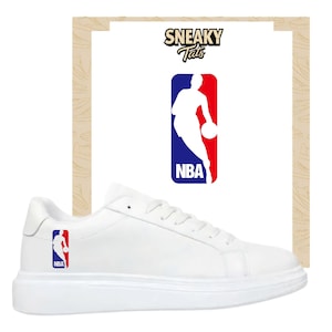 Puede incluir: Zapatillas blancas con el logo de la NBA en rojo, blanco y azul. El logo también aparece sobre un fondo blanco con el texto "Sneaky Tats". Las zapatillas tienen una suela blanca gruesa.