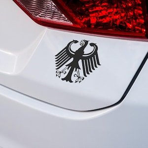 Op de afbeelding: Zwarte sticker van een gestileerde adelaar, een symbool van Duitsland, bevestigd op een witte auto. De adelaar heeft uitgespreide vleugels en is gecentreerd op het oppervlak van het voertuig. Het rode achterlicht van de auto is zichtbaar op de achtergrond.