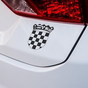 Könnte beinhalten: Ein schwarz-weiß kariertes Emblem mit Kronenmotiv, befestigt an einem weißen Auto. Das Emblem hat die Form eines Schildes, mit einem Schachbrettmuster und einer detaillierten Krone oben. Das rote Rücklicht des Autos ist im Hintergrund sichtbar.
