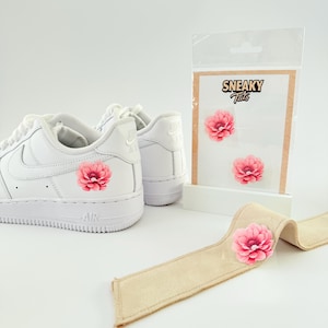 May include: White trainers with pink flower decals. A package of flower decals is displayed next to the shoes. A tan fabric strip with a pink flower decal is in the foreground. The package has the text "Sneaky Tats".