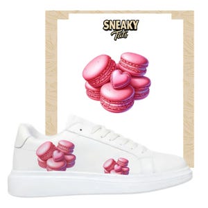 Peut inclure: Baskets blanches avec des graphiques de macarons roses. Les chaussures présentent un groupe de macarons sur le côté et un plus grand graphique de macarons avec un macaron en forme de cœur sur la languette. Le texte "Sneaky Tats" est en haut.