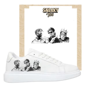 Peut inclure: Baskets blanches avec un graphisme noir et blanc de trois rappeurs célèbres. Les baskets ont un profil bas et une semelle blanche. Le texte "Sneaky Tats" est au-dessus de l'image des rappeurs.