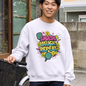 Puede incluir: Sudadera blanca con un gráfico colorido que dice "SMASH, LAUGH, REPEAT" con palas de pickleball y caras sonrientes. El diseño presenta colores rosa, azul, amarillo y verde. La sudadera es de estilo casual.
