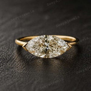 Op de afbeelding: Een gouden ring met een marquise-geslepen diamant. De diamant heeft een lichte champagnekleur en is gezet in een eenvoudige band. De ring wordt getoond op een donkere ondergrond, waardoor de glans en het ontwerp worden benadrukt.