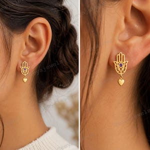 May include: Gold-coloured Hamsa hand earrings, each featuring a small purple gemstone and a heart charm. These delicate earrings are designed for everyday wear. The Hamsa hand is a symbol of protection and good fortune, crafted in a fine gold finish.