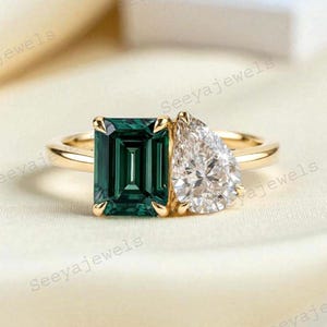May include: A gold ring featuring two gemstones: a rectangular emerald-cut green stone and a pear-shaped white stone. The ring is set on a cream-colored fabric, with the brand name "Seeyajewels" visible in the background.