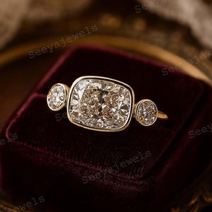 Puede incluir: Un anillo de oro con un gran diamante central de talla cuadrada y dos diamantes redondos más pequeños a cada lado. El anillo está sobre una superficie de terciopelo rojo oscuro. Es una joya.