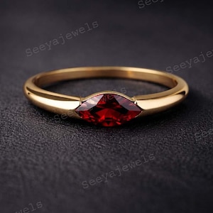 Puede incluir: Un anillo de oro con una piedra preciosa roja oscura con corte marquesa. El anillo tiene una banda lisa y pulida y un engaste que sujeta de forma segura la piedra preciosa. El anillo se muestra sobre una superficie oscura.
